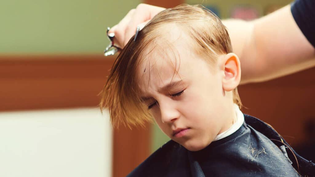 Cute little boy getting haircut by hairdresser at the barbershop. Barber man doing kid the hairstyle.